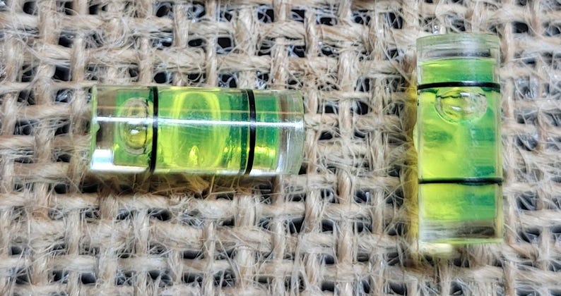 May include: Two clear plastic spirit levels with green liquid and black lines. The levels are on a brown burlap background.