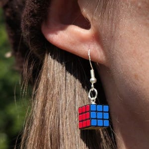 May include: A dangling earring featuring a miniature cube puzzle charm with red, blue, and yellow grid-patterned sides. The charm is attached to a silver-toned metal hook wire, designed to hang from an earlobe with a classic fishhook style.