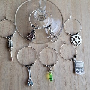 May include: Set of six silver wine glass charms with different charms, including a robot, lightbulb, wrench, screwdriver, level, and laptop.