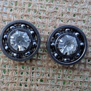 May include: Two silver-toned, round earrings with a central, faceted, clear crystal. The earrings have a ball bearing design around the crystal. The background is a rough, woven, tan fabric.