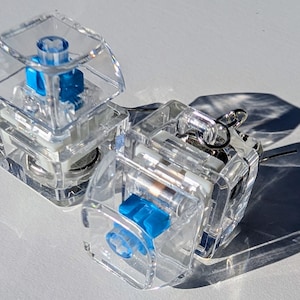 May include: Three clear mechanical keyboard switches with blue accents. The switches are transparent, allowing a view of the internal components. The blue components add a pop of color. The switches are designed for use in keyboards.