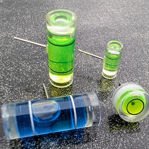 May include: Four small level tools with green and blue liquid inside. The level tools are made of clear glass and have a metal pin on one end. The level tools are on a gray surface.