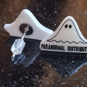 May include: A pair of white ghost-shaped earrings with black text that reads "Paranormal Distribution".