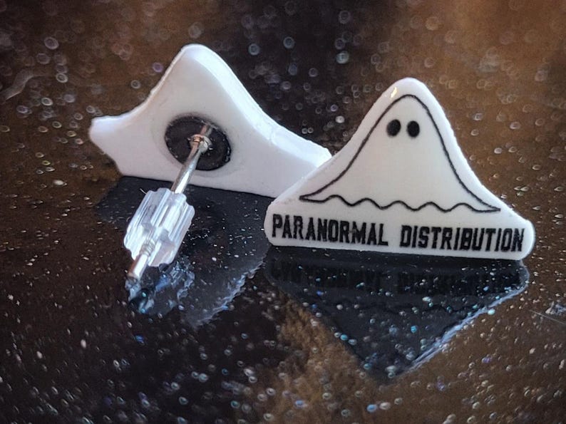 May include: A pair of white ghost-shaped earrings with black text that reads "Paranormal Distribution".