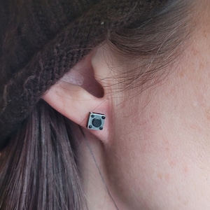 May include: A pair of silver square stud earrings with a black center button. The earrings are worn in a person's ear.