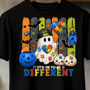 May include: A black t-shirt featuring a graphic of a ghost wearing a baseball cap, holding a heart-shaped puzzle piece balloon. The design includes a blue pumpkin bucket, two small dumplings, bats, and the text IT'S OK TO BE DIFFERENT in colorful letters.