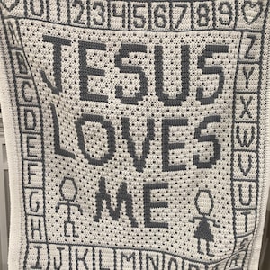 May include: A white crocheted blanket with the words "JESUS LOVES ME" in gray yarn. The blanket also features a gray alphabet border and a gray stick figure of a person on each side of the words.
