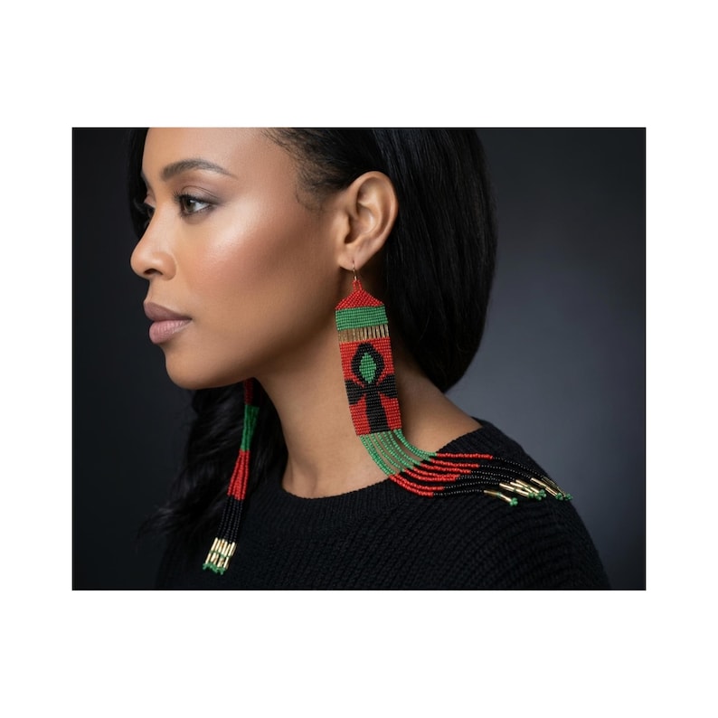 Puede incluir: Pendientes largos de cuentas en rojo, verde, negro y dorado. Los pendientes presentan un s&iacute;mbolo Ankh y un dise&ntilde;o de flecos. Los pendientes los lleva una persona con cabello oscuro y una parte superior negra.