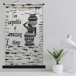 May include: Black and white wall hanging with the text "I am capable of amazing things" and a silhouette of a woman carrying a pot on her head. The background has a textured, distressed look.