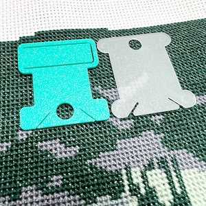 Op de afbeelding: Twee plastic flossspoelen, &eacute;&eacute;n turquoise en &eacute;&eacute;n doorschijnend, liggen op een kruissteekproject. De turquoise spoel heeft een rechthoekige bovenkant met een gat en een sleuf voor draad. De doorschijnende spoel heeft een vergelijkbare vorm met een gat.