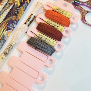 Peut inclure: Un ensemble de fournitures de broderie comprenant un morceau de toile à broder, un écheveau de fil crème et quatre couleurs de fil à broder enroulées sur des bobines en plastique rose clair étiquetées 891, 902, 924 et 948, avec des ciseaux en métal irisé.