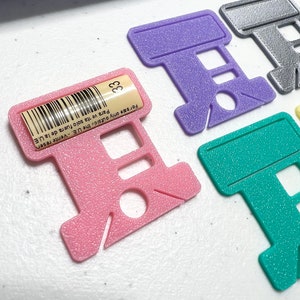 Op de afbeelding: Een verzameling gekleurde plastic draadorganizers in roze, paars, teal en zilver. De roze organizer heeft een label met nummer 33 en tekst. Deze organizers zijn voor kruissteek- of borduurgaren.