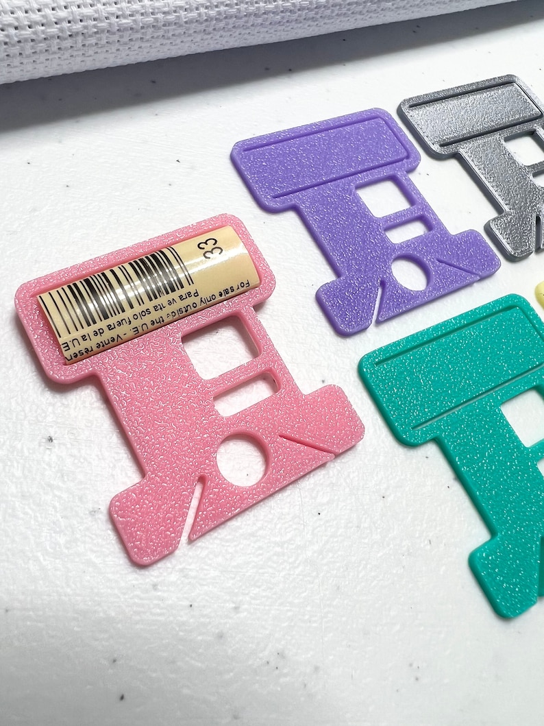 Op de afbeelding: Een verzameling gekleurde plastic draadorganizers in roze, paars, teal en zilver. De roze organizer heeft een label met nummer 33 en tekst. Deze organizers zijn voor kruissteek- of borduurgaren.