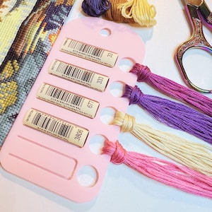 May include: A pink plastic embroidery floss organiser card holding four strands of thread in shades of magenta, cream, purple, and dark pink. Each thread is labelled with a barcode and colour number. Nearby are three loose thread bundles and scissors.