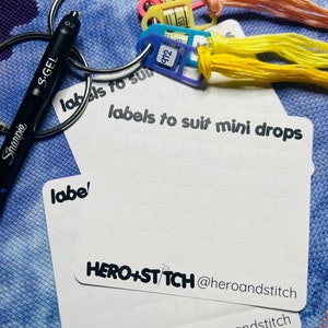 Könnte beinhalten: Zwei weiße Blätter Papier mit einem Raster aus Quadraten zum Beschriften von Stickgarn. Die Blätter tragen den Text "labels to suit mini drops" und "HERO+ST/TCH @heroandstitch". Es gibt drei Plastik-Garnrollen mit Nummern darauf und einen schwarzen Sharpie-Marker.