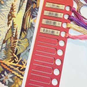 May include: A red plastic embroidery floss organizer with multiple slots and holes, holding four strands of colored thread in purple, light yellow, and pink. Next to it is a piece of cross-stitch fabric with a landscape pattern and metal scissors.