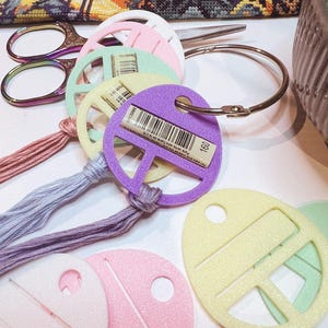 May include: A collection of pastel-coloured plastic embroidery floss bobbins in shades of pink, yellow, green, and purple, organised on a silver metal ring. Each bobbin features a barcode label and is attached to a matching strand of embroidery thread.