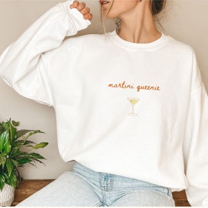 Peut inclure: Sweat-shirt blanc avec le texte "martini queenie" en écriture orange et une petite illustration d'un verre à martini avec une olive.