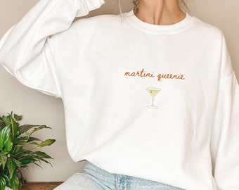 May include: White sweatshirt with the text "martini queenie" in orange script and a small illustration of a martini glass with an olive.