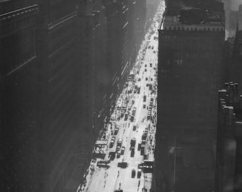 May include: A black and white aerial photograph of a busy city street lined with tall buildings. The street is filled with cars and pedestrians.