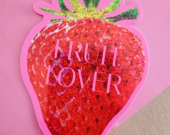 Può includere: Un adesivo a forma di fragola rossa con glitter con il testo "FRUIT LOVER" in lettere bianche.
