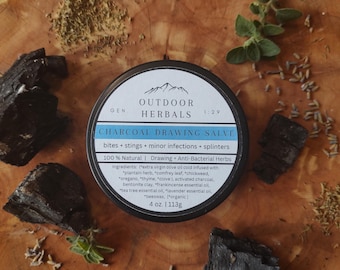 Op de afbeelding: Een ronde blik met houtskooltekenzalf, omringd door stukjes houtskool en kruiden, staat op een houten oppervlak. Het etiket luidt "Outdoor Herbals Charcoal Drawing Salve" en vermeldt ingrediënten en toepassingen. De blik bevat 113g.