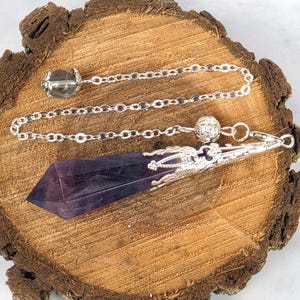 Peut inclure: Un pendentif pendule en cristal violet à facettes et pointu, doté d'un capuchon en métal argenté avec des détails en filigrane complexes. Le pendentif est fixé à une chaîne en métal argenté comprenant une petite perle argentée texturée.