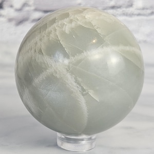 May include: A smooth, white and grey moonstone sphere on a clear acrylic stand. The sphere has a natural, iridescent sheen.