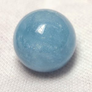 Può includere: Una pietra preziosa lucida e sferica di colore azzurro traslucido. La superficie della pietra è liscia e riflettente, mostrando sottili texture interne e variazioni nella tonalità blu su tutta la forma arrotondata dell'oggetto.