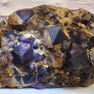 May include: A cluster of deep purple, translucent fluorite crystals embedded in a textured, tan-coloured rocky matrix. The mineral specimen features numerous sharp, geometric cubic and octahedral crystal formations with varying levels of transparency.