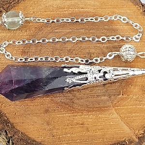 May include: A pointed, deep purple amethyst crystal pendulum encased in a decorative silver-toned metal cap. The crystal is attached to a silver-toned metal chain featuring a small, textured silver bead and a pale green translucent bead at the end.