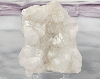 Pode incluir: Um aglomerado de cristais de quartzo branco translúcido com facetas afiadas e angulares e uma superfície brilhante e reflexiva. A formação mineral está montada num suporte de exibição de acrílico transparente.