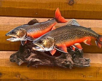 May include: A decorative wall hanging featuring two detailed fish sculptures. The fish have orange and red fins and tails, with speckled bodies. They are mounted on a piece of driftwood against a wooden background.