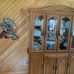 May include: A wooden wall hanging featuring three crappie fish sculptures mounted on driftwood. The fish are painted with realistic details and colours. A large wooden cabinet with glass doors is in the background, displaying dishes and glassware.