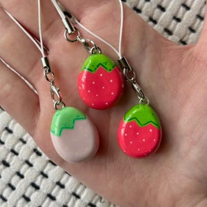 May include: Three strawberry-shaped charms with white cords and silver-tone clasps. Two charms are red with white dots and green tops. The third charm is pink with white dots and a green top. The charms are held in a hand.