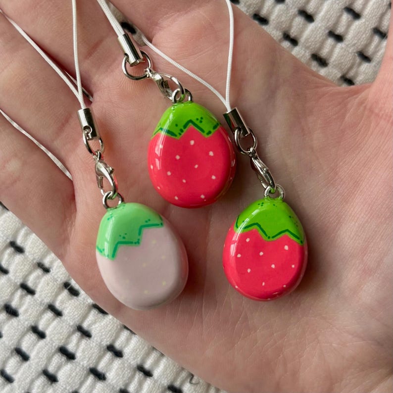 May include: Three strawberry-shaped charms with white cords and silver-tone clasps. Two charms are red with white dots and green tops. The third charm is pink with white dots and a green top. The charms are held in a hand.