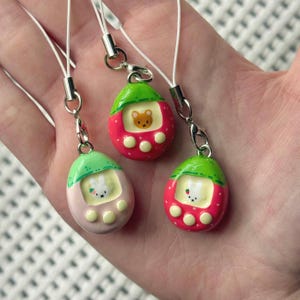 May include: Three colorful, handmade strawberry-shaped charms with white cords. Each charm features a different animal character on a screen: a bear, a bunny, and a cat. The charms are red, pink, and green with white accents.