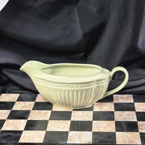 Könnte beinhalten: Eine hellgrüne Sauciere mit Henkel und Ausguss. Das Gefäß hat ein geripptes Design am unteren Teil und einen dekorativen Rand am Rand. Die Sauciere ist aus Keramik oder Porzellan gefertigt und wahrscheinlich vintage.