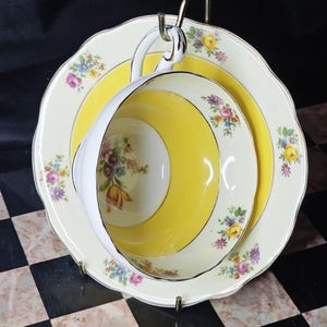 May include: A vintage teacup and saucer set with a yellow and white design. The teacup and saucer have floral accents and gold trim. The set is displayed on a gold-coloured metal stand. The teacup has a white handle.