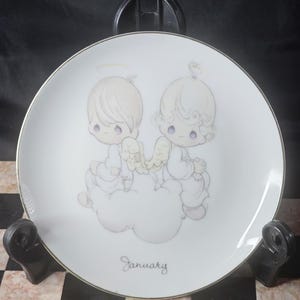 May include: A white decorative plate with a gold rim featuring two cherubic figures with halos and wings, seated on a cloud. The word "January" is written in script below the figures.