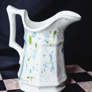 May include: White ceramic jug with a handle and spout. The jug is decorated with blue and green speckled paint. The jug is sitting on a black and white checkered surface.