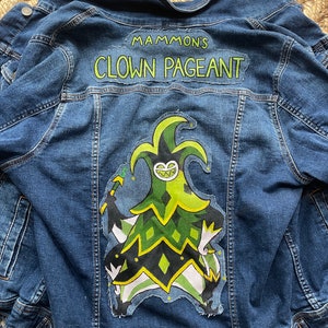 May include: A blue denim jacket with a large patch on the back featuring the text "MAMMON'S CLOWN PAGEANT" above a cartoon jester character in green, black, and white. The jacket has button closures and pockets.
