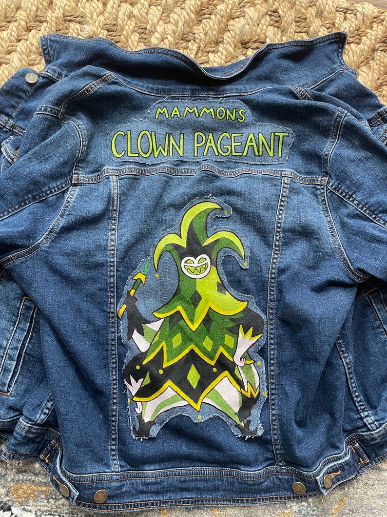 May include: A blue denim jacket with a large patch on the back featuring the text "MAMMON'S CLOWN PAGEANT" above a cartoon jester character in green, black, and white. The jacket has button closures and pockets.