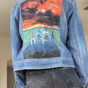 May include: A denim jacket with a painted scene on the back featuring a sunset and figures on bicycles. The artwork includes a road, green grass, and a sign that reads "Hawkins". The jacket is a medium wash blue.