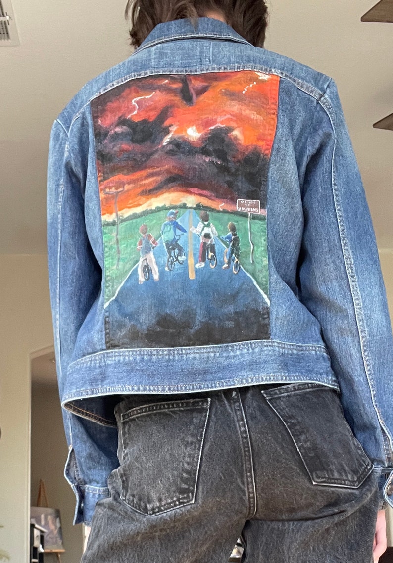 May include: A denim jacket with a painted scene on the back featuring a sunset and figures on bicycles. The artwork includes a road, green grass, and a sign that reads "Hawkins". The jacket is a medium wash blue.