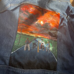 May include: Blue denim jacket with a painted scene on the back. The artwork depicts four figures on bicycles on a road under a sunset sky. The scene includes a sign that reads "Hawkins".