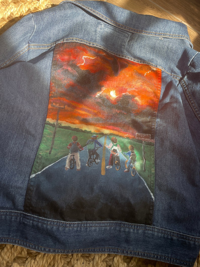 May include: Blue denim jacket with a painted scene on the back. The artwork depicts four figures on bicycles on a road under a sunset sky. The scene includes a sign that reads "Hawkins".