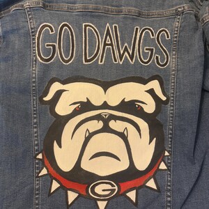 May include: Blue denim jacket with the words "GO DAWGS" in white lettering. Below the text is a bulldog graphic with a red collar and the letter "G". The jacket is a medium wash denim.