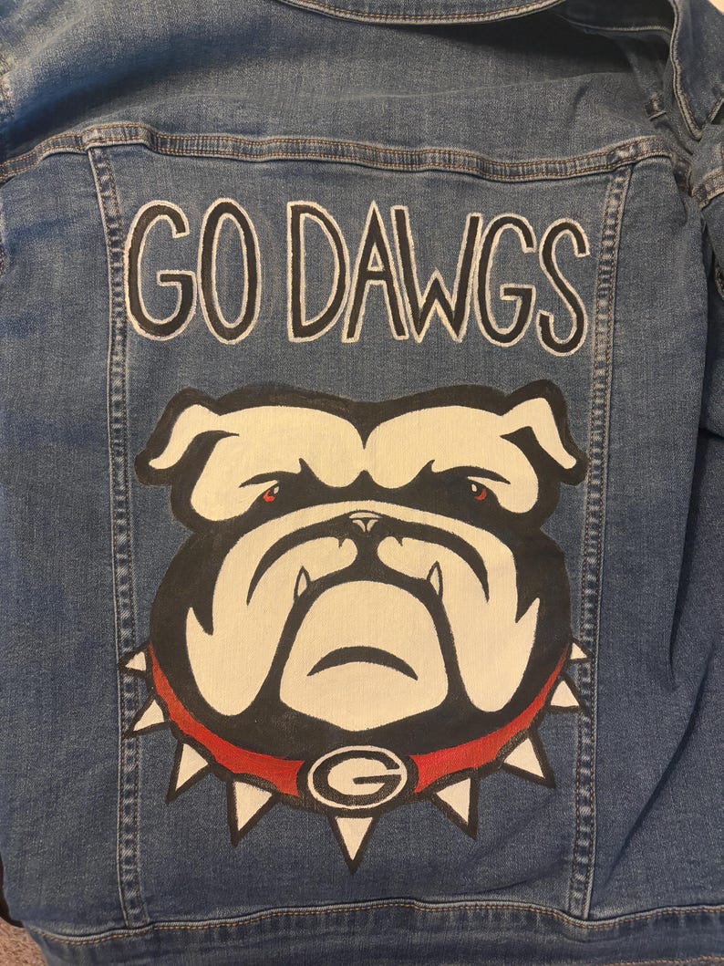 May include: Blue denim jacket with the words "GO DAWGS" in white lettering. Below the text is a bulldog graphic with a red collar and the letter "G". The jacket is a medium wash denim.