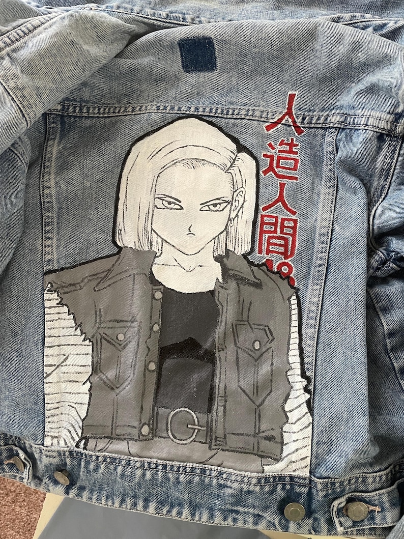 May include: Light blue denim jacket featuring a hand-painted illustration of an anime character. The artwork depicts a woman with short blonde hair and a black and gray outfit. Japanese text is visible on the upper right side of the jacket.